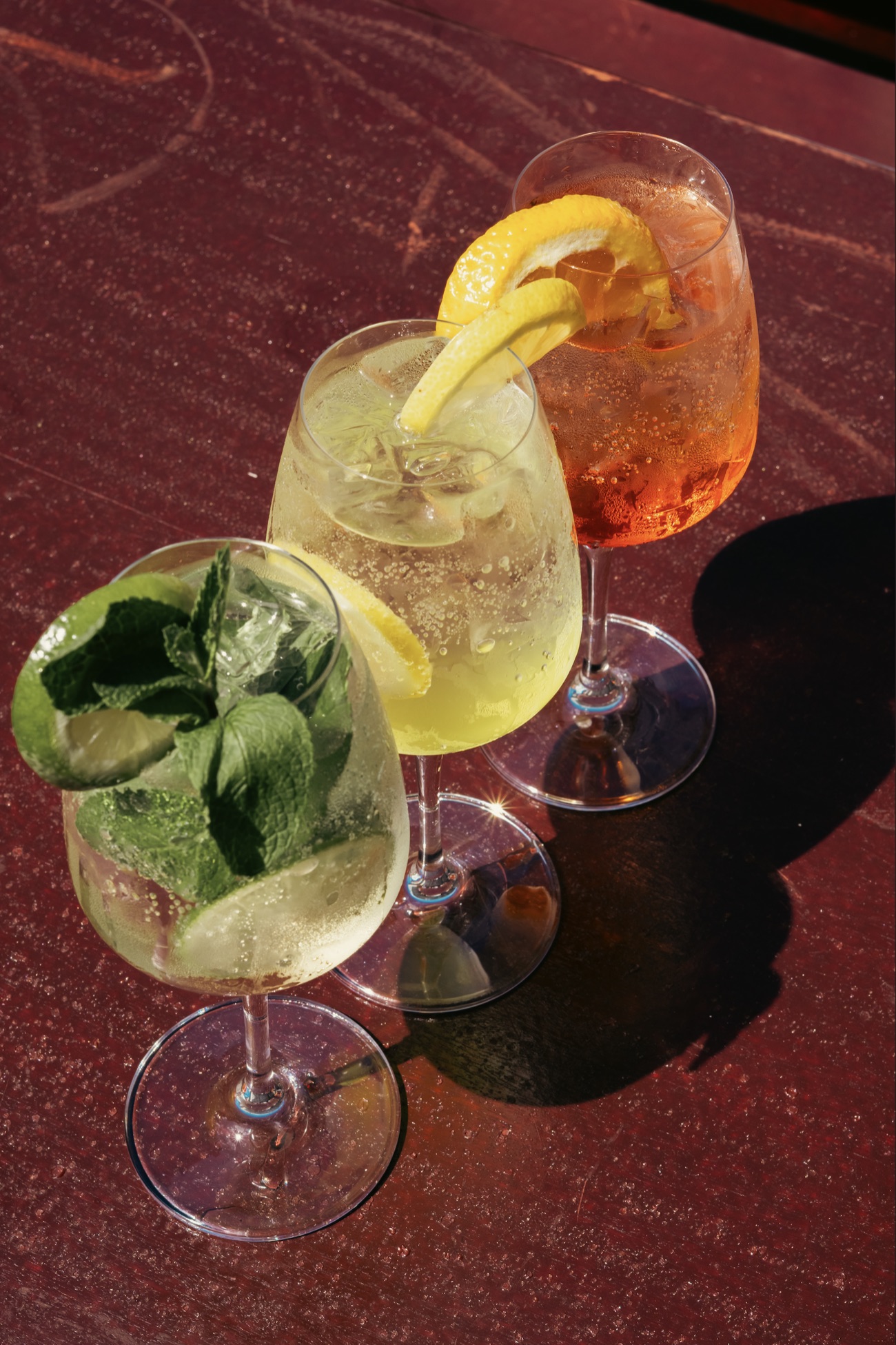 Three spritz cocktails garnished with mint, lemon and orange on a mahogany table